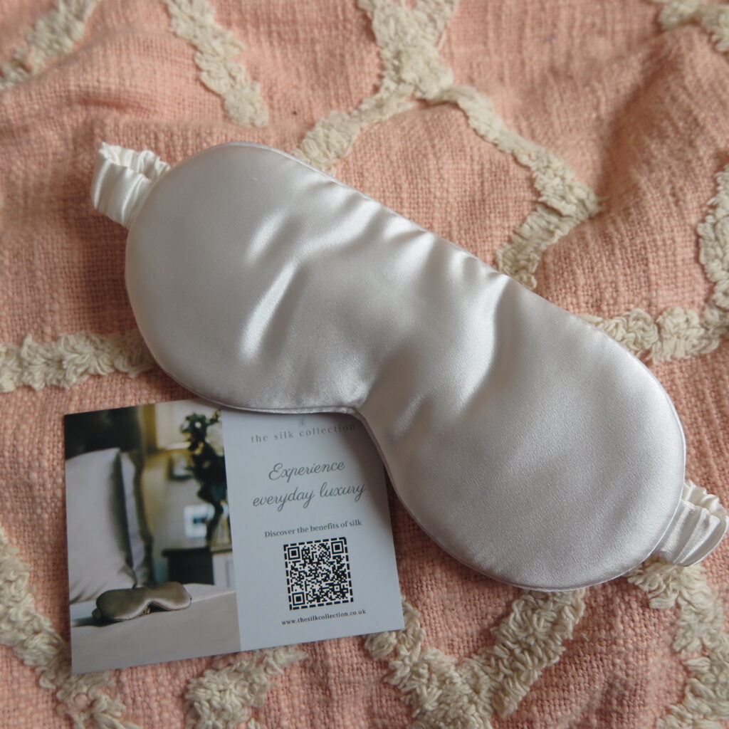 Image of The Silk Collection eye mask along with information card on a pink bedspread.