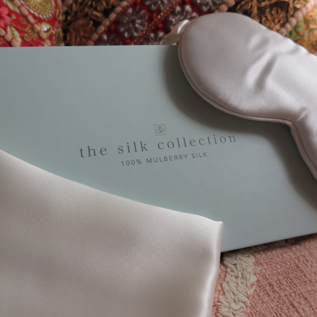 Image of The Silk Collection mask in pearl white next to the presentation box and a silk pillowcase
