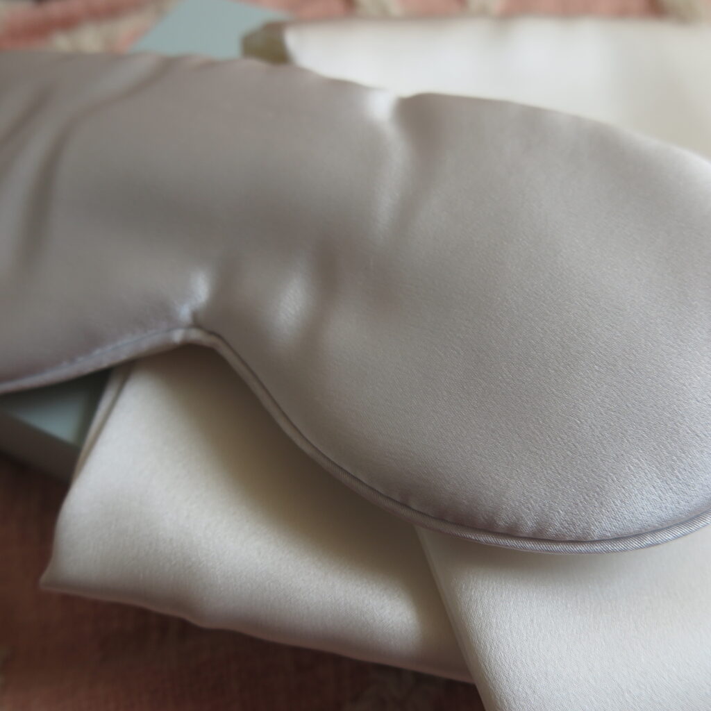 Close up image of the silk eye mask laid on top of a silk pillowcase, detailing the stitching and lustre of the product