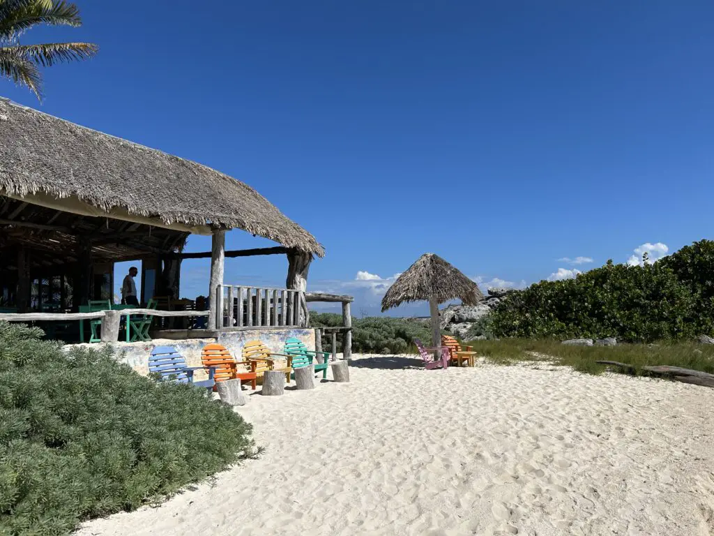 A photograph of the Restaurant palapa at Zamas Hotel under a background of blue skies. Colourful 'Sunrise style' chairs feature in the mid-ground, on the golden sands of the foreground.